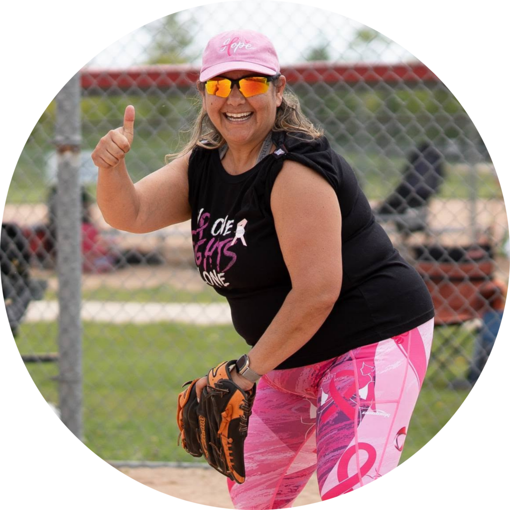 A woman wearing a cancer survivor hat and baseball glove gives a thumbs up while on the diamond.