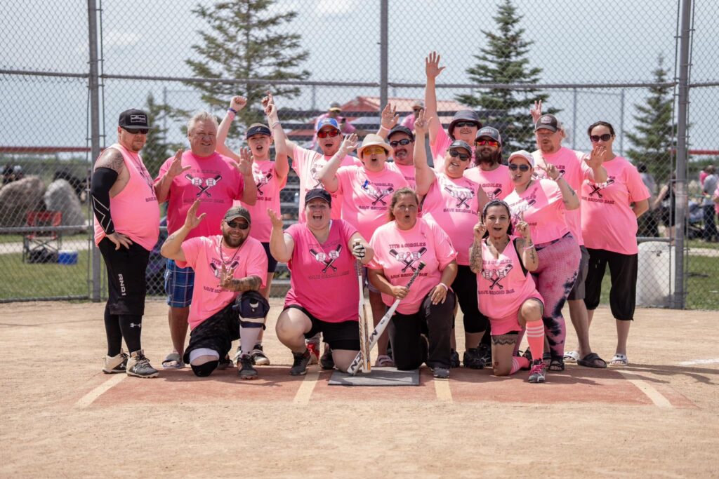 A baseball team (Tits & Giggles) pose for a photo at home plate on the diamond.
