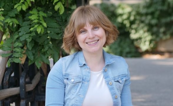 A woman with short strawberry blonde hair sits on the ground outside smiling.